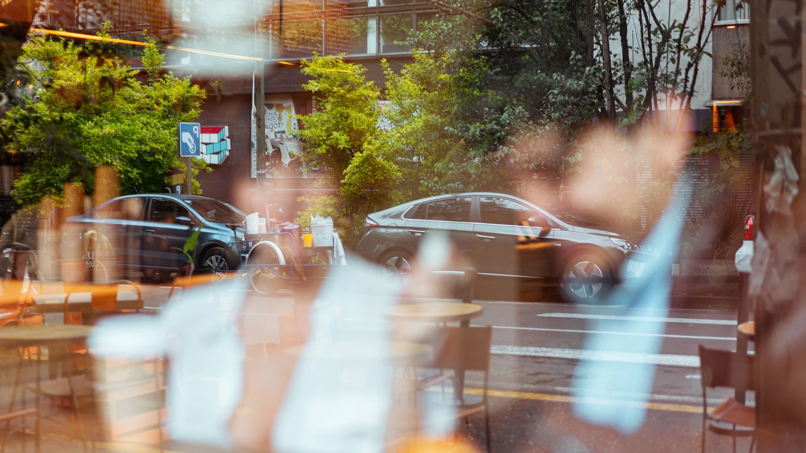 Riflessi della vista della strada attraverso il vetro di un caffè che mostra auto parcheggiate e alberi verdi.