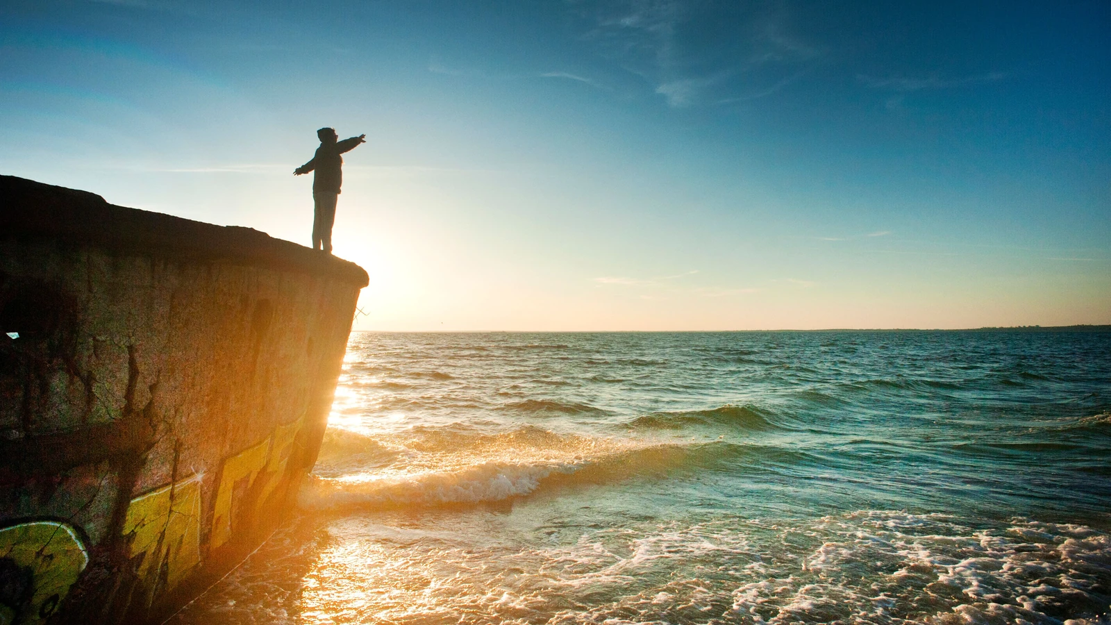 Una persona su una rupe guarda l'oceano al tramonto con le braccia sollevate in un momento di gioia e riflessione.