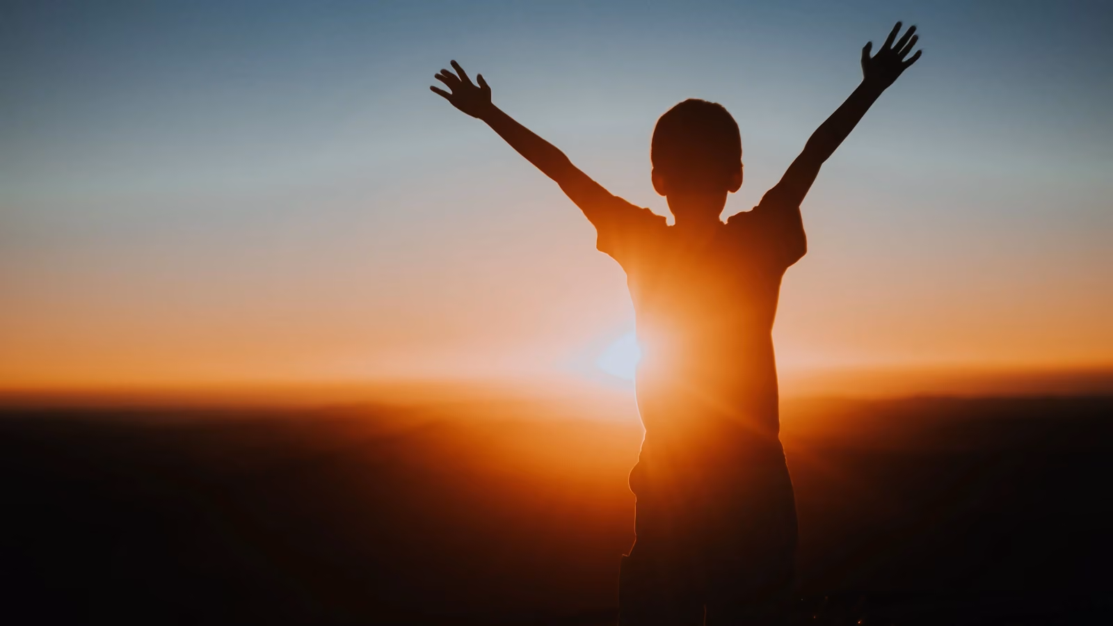 Silhouette di un bambino con le braccia alzate che gode di un vibrante tramonto, simboleggiando libertà e gioia. — Matheus Bertelli – Pexels
