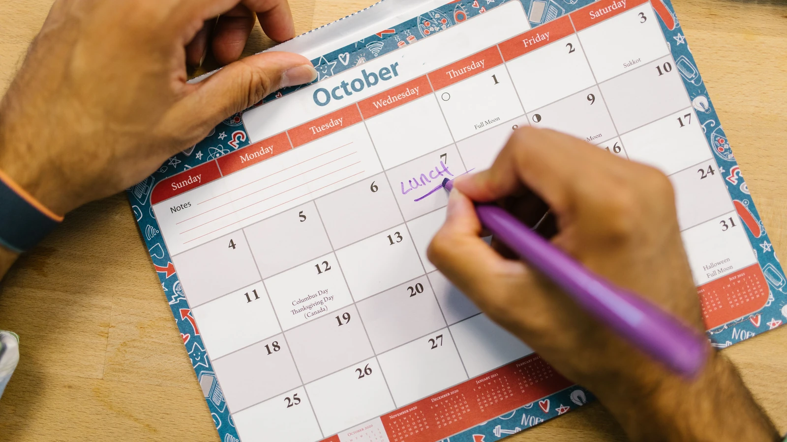 Primo piano di una persona che scrive un promemoria per il pranzo su un calendario di ottobre con una penna viola.