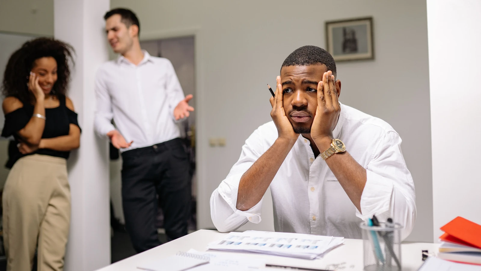 Un dipendente stressato è seduto a una scrivania con colleghi che discutono nelle vicinanze, evidenziando la tensione sul posto di lavoro. — Yan Krukau – Pexels