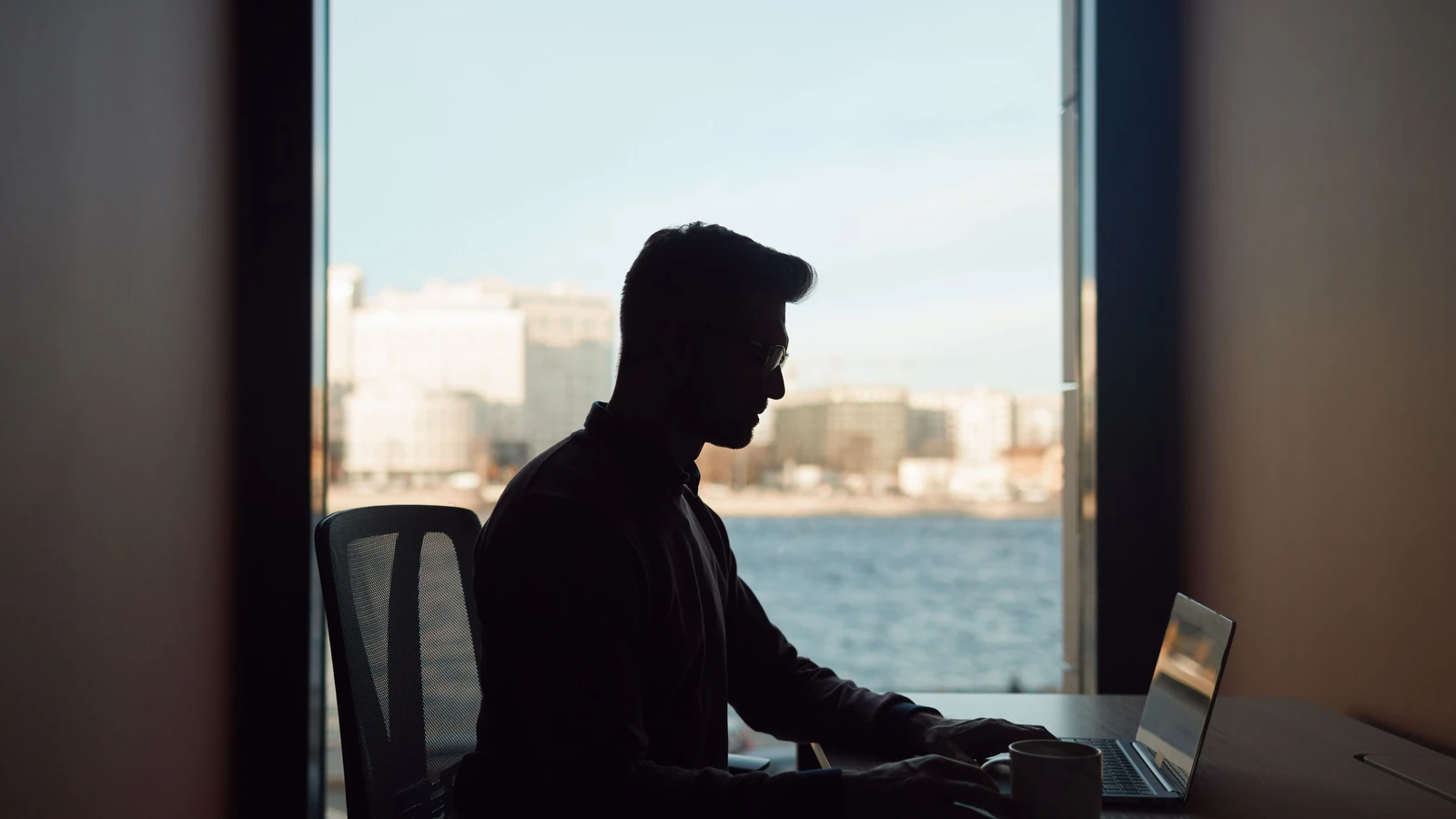 Silhouette di un uomo che lavora al suo laptop accanto a una finestra con vista su un paesaggio urbano costiero.
