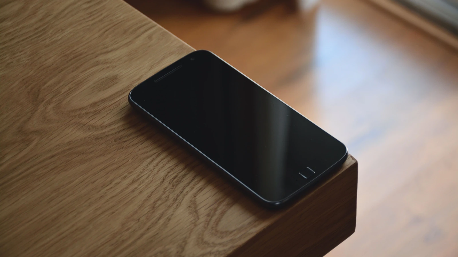 Uno smartphone elegante si appoggia sul bordo di un tavolo in legno in un ambiente interno illuminato caldamente.