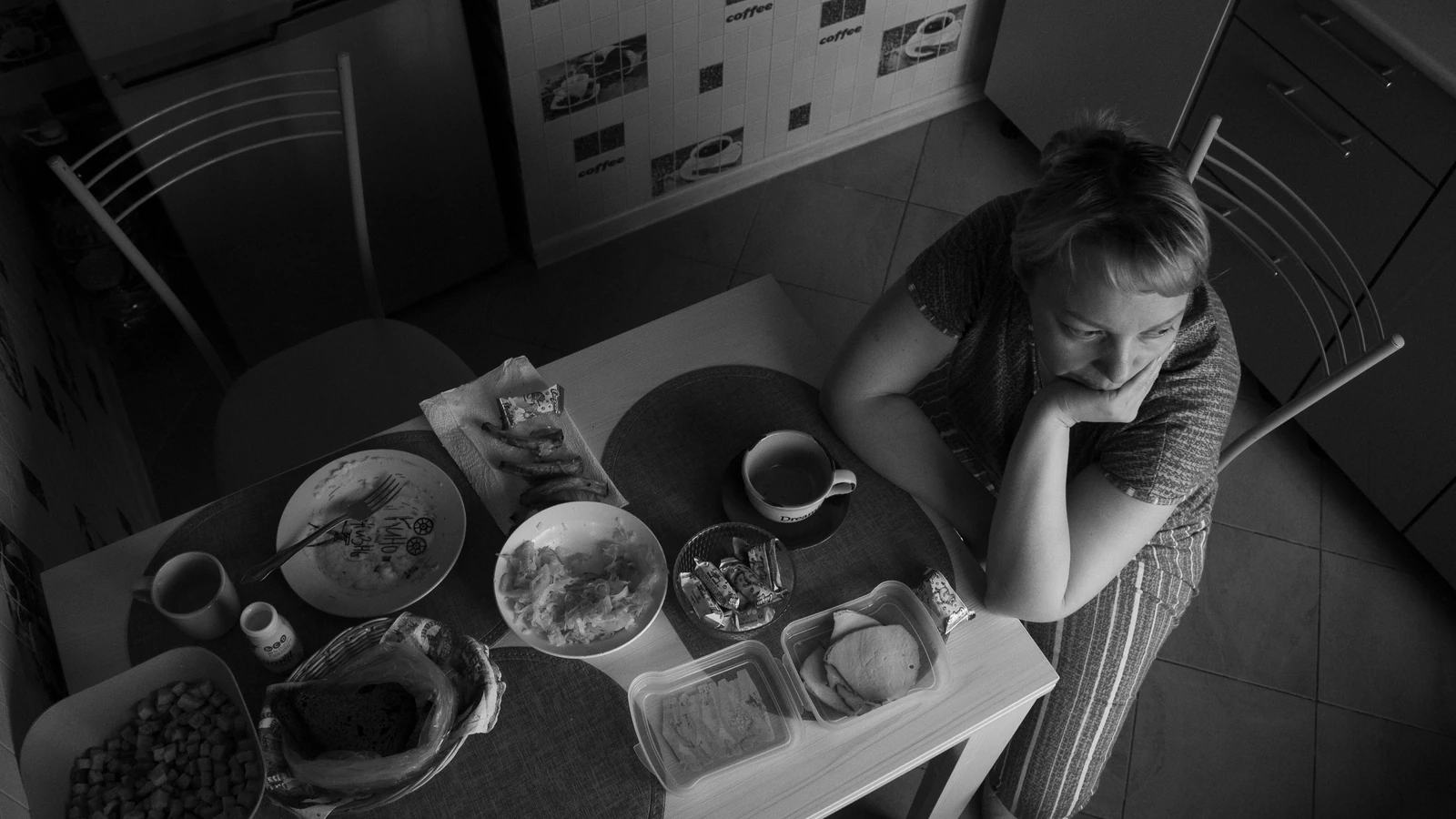 Vista in bianco e nero di una donna seduta a un tavolo di cucina con articoli per la colazione.