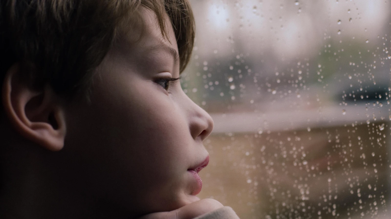 Bambino contemplativo scruta attraverso la finestra spruzzata di pioggia, evocando un senso di calma e riflessione.
