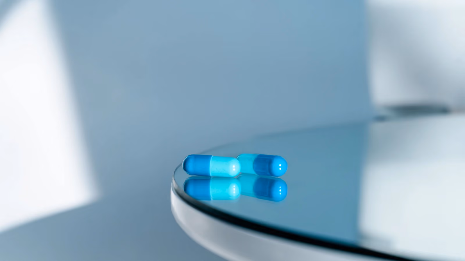Primo piano di due capsule blu su un tavolo rotondo di vetro lucido, che riflettono la luce. — Natalie Bond – Pexels