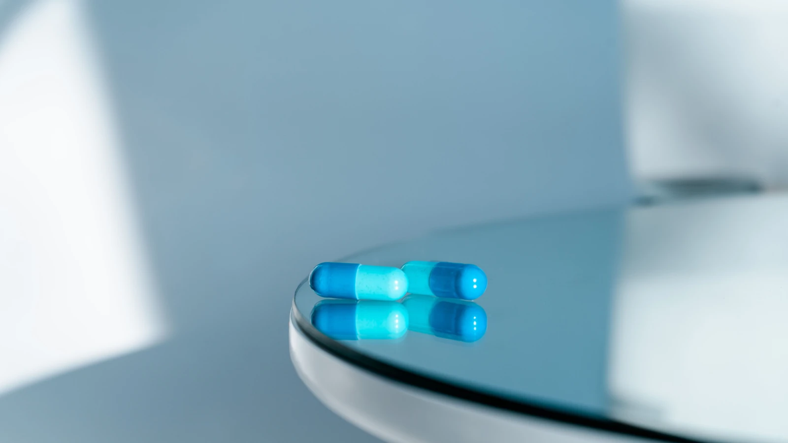Primo piano di due capsule blu su un tavolo rotondo di vetro lucido, che riflettono la luce. — Natalie Bond – Pexels
