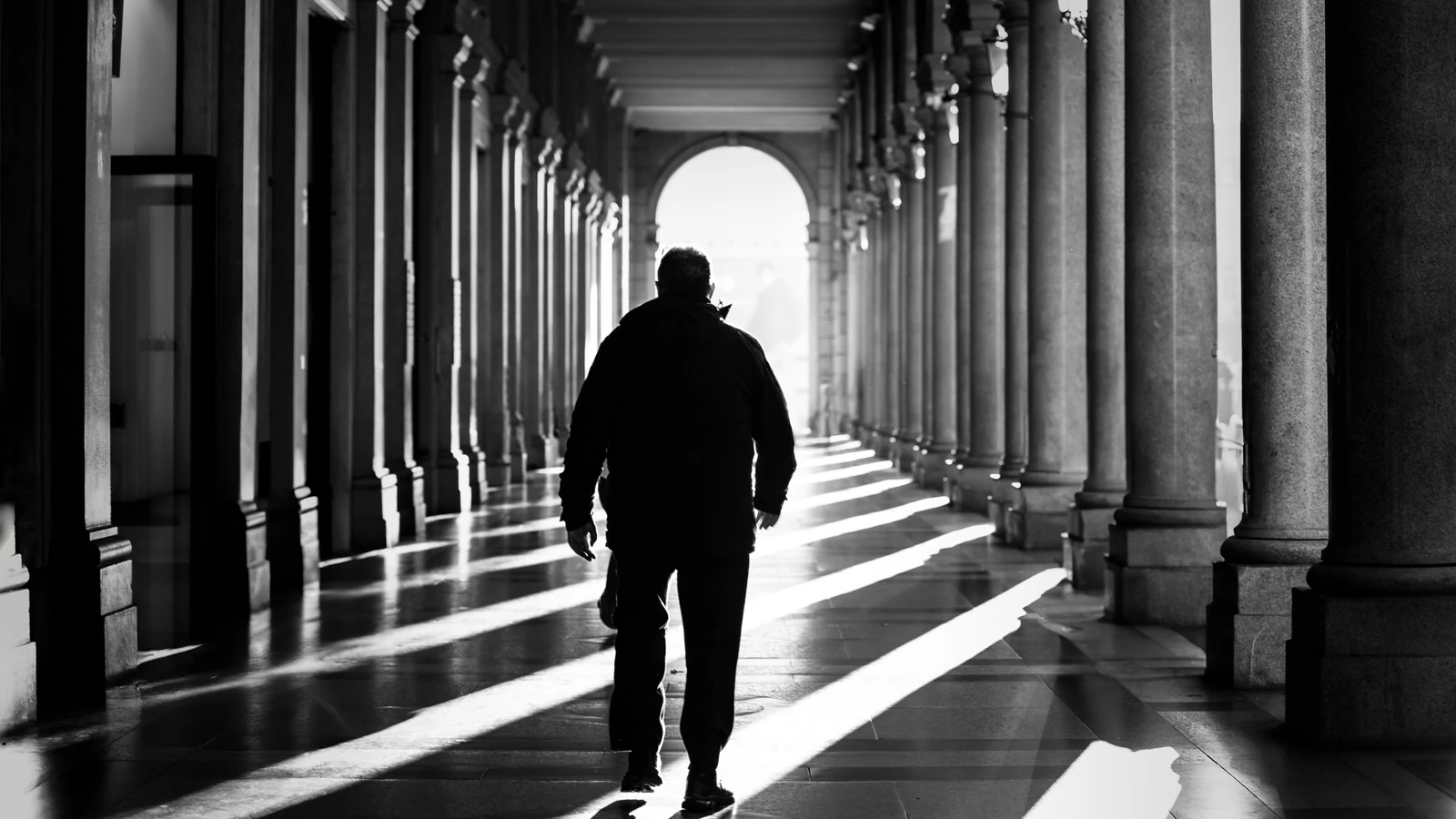 Silhouette di un uomo che cammina attraverso una colonnata illuminata dal sole a Torino, Italia, catturata in straordinario bianco e nero.