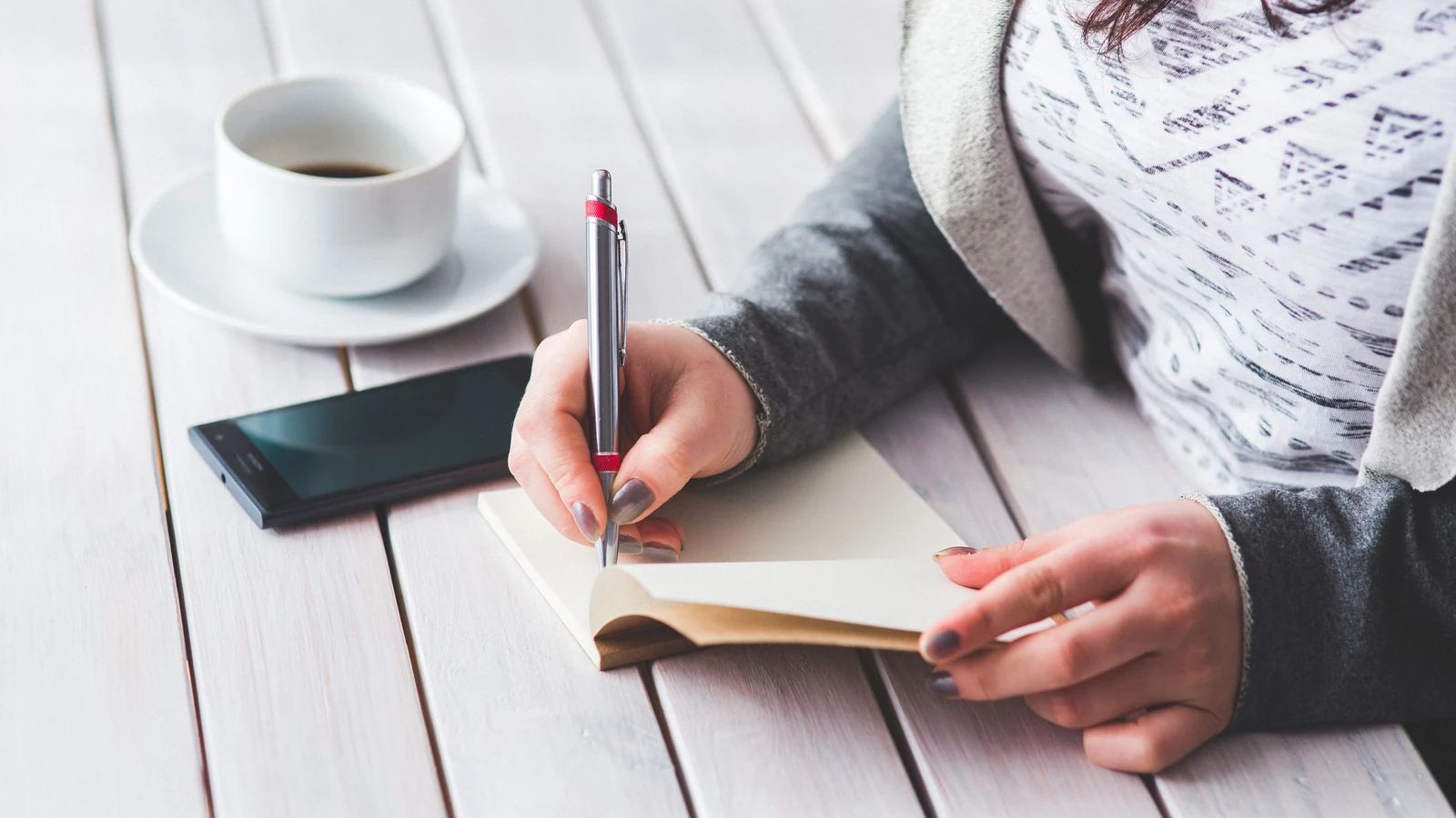 Una donna scrive in un diario accanto a una tazza di caffè e uno smartphone su un tavolo di legno.