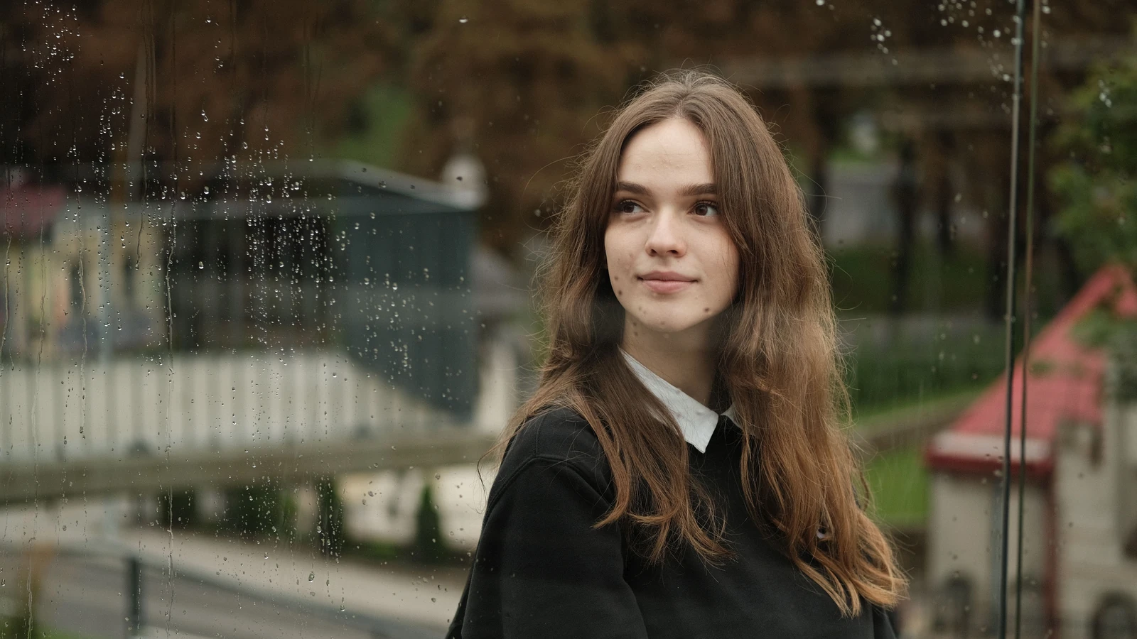 Una giovane donna con lunghi capelli castani guarda pensierosa attraverso una finestra spruzzata di pioggia in un contesto urbano.
