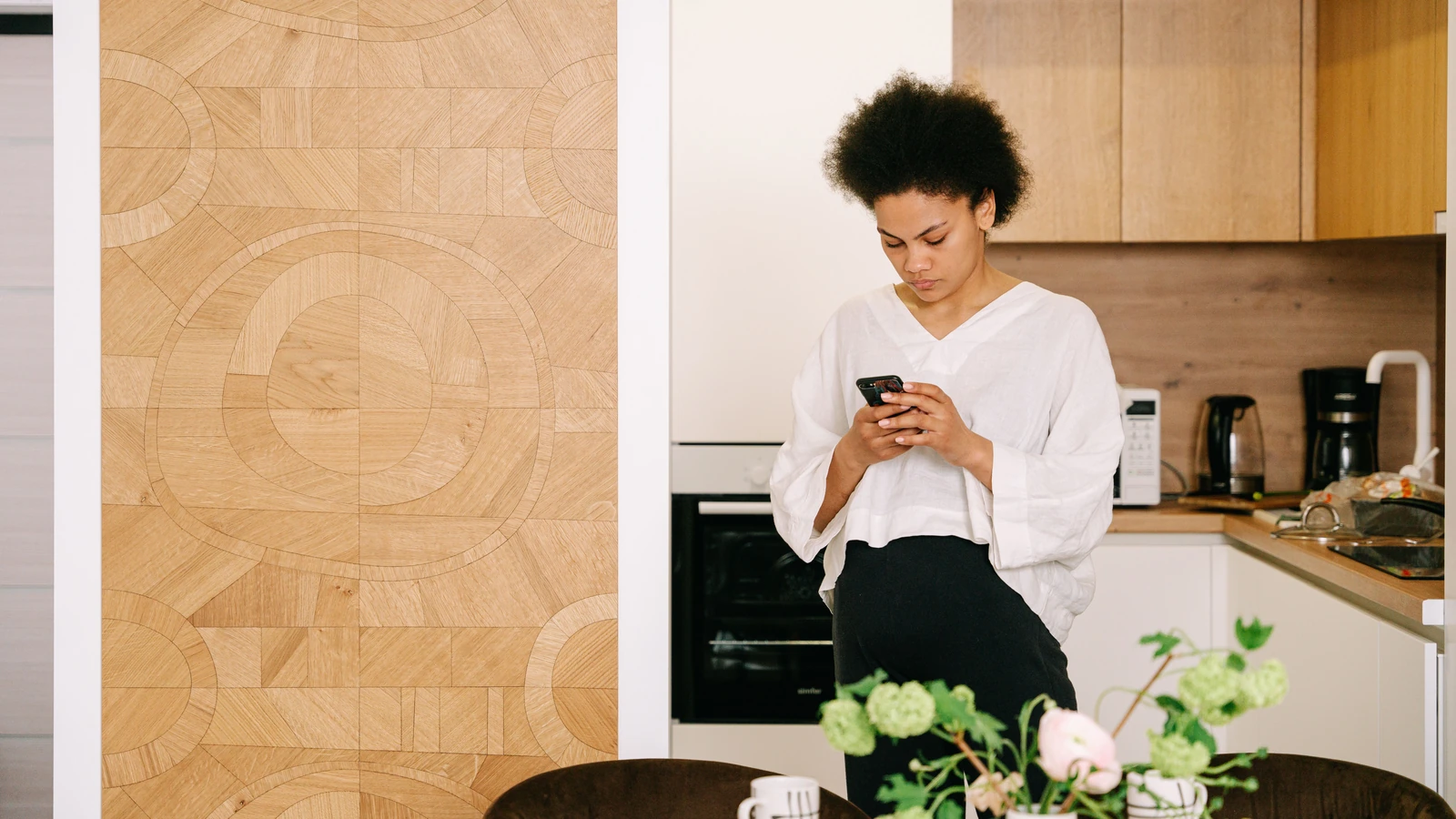 Donna afroamericana con capelli afro che invia messaggi su un cellulare in una cucina moderna e accogliente.