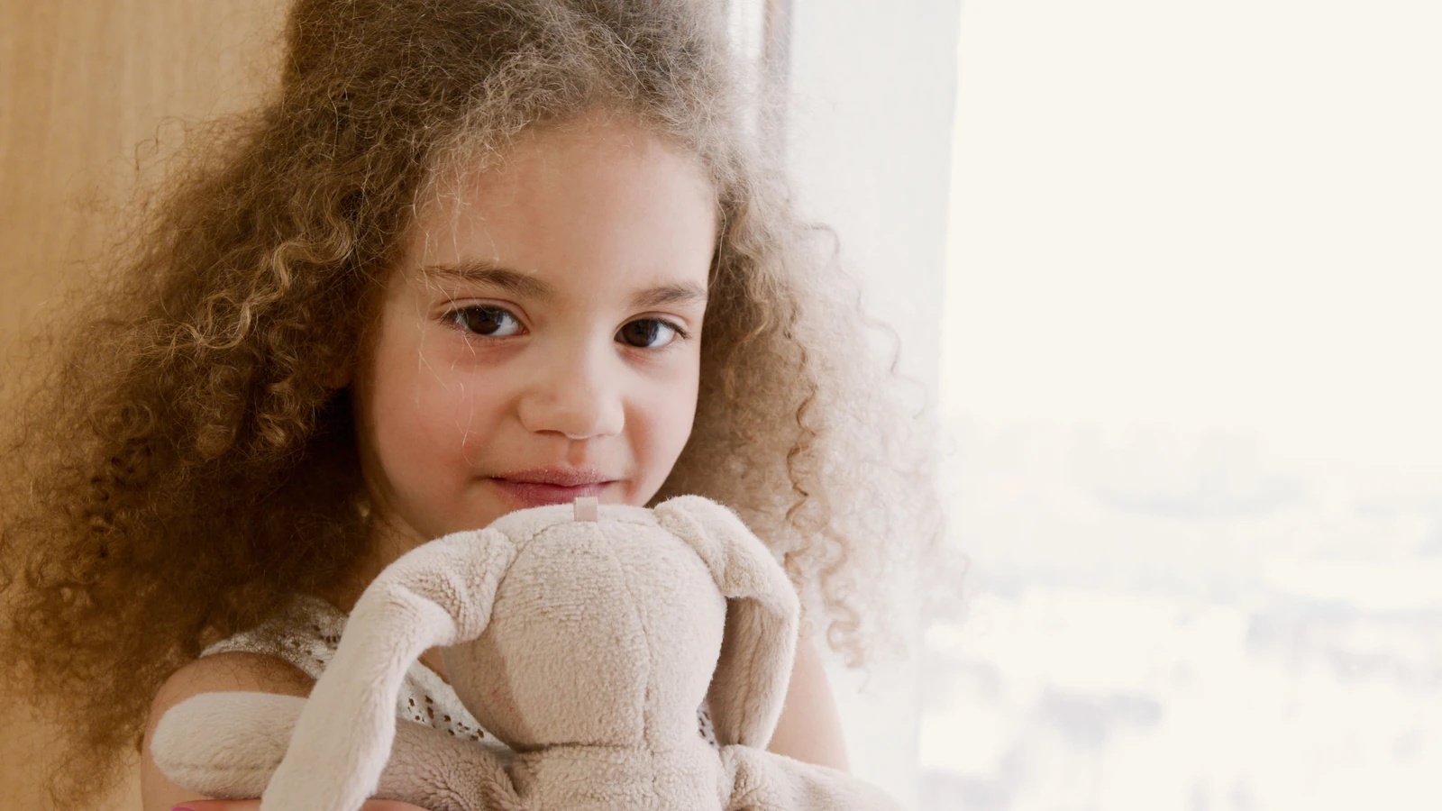 Adorabile bambina con capelli ricci che tiene un coniglio di peluche in casa, sorridendo dolcemente.