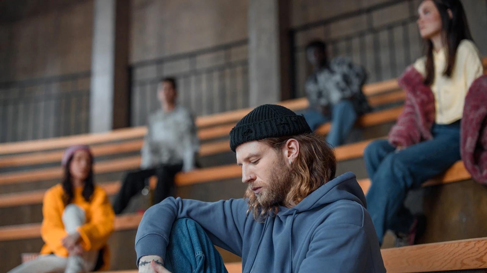 Un gruppo diversificato di adulti seduti in un auditorio, concentrati su un uomo pensieroso con felpa e cappello di lana. — Ron Lach – Pexels
