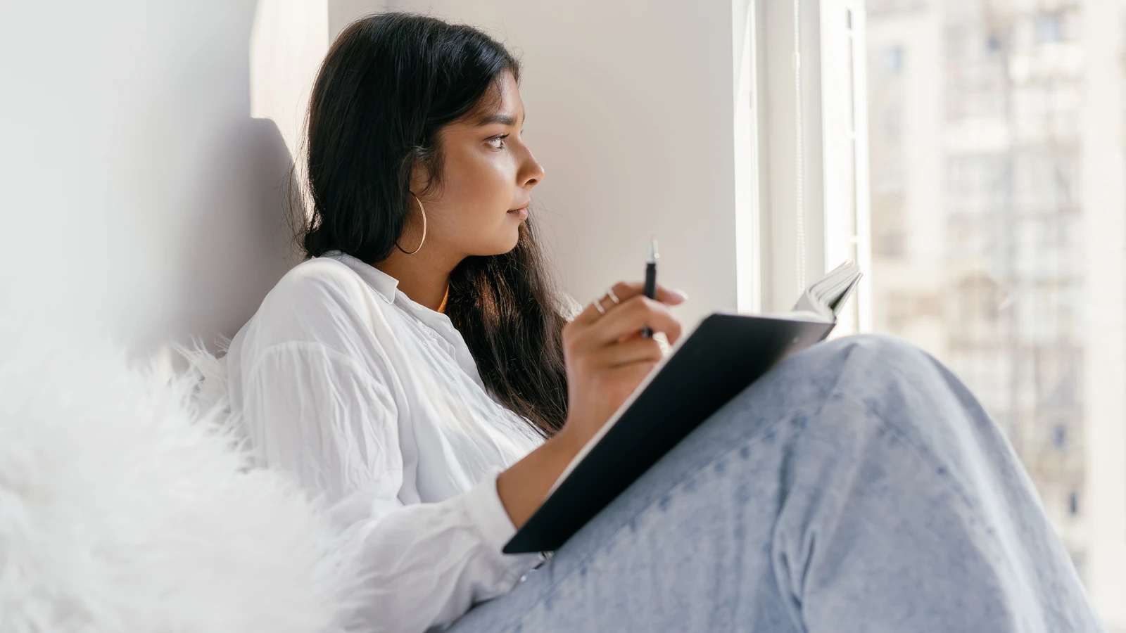 Una giovane donna seduta accanto a una finestra, che studia pensierosa con un quaderno in mano. Ambiente di apprendimento interno.