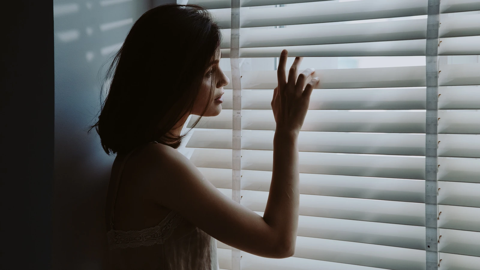 Una giovane donna in camicia da notte guarda pensierosa attraverso le persiane, creando un'atmosfera serena e introspettiva.