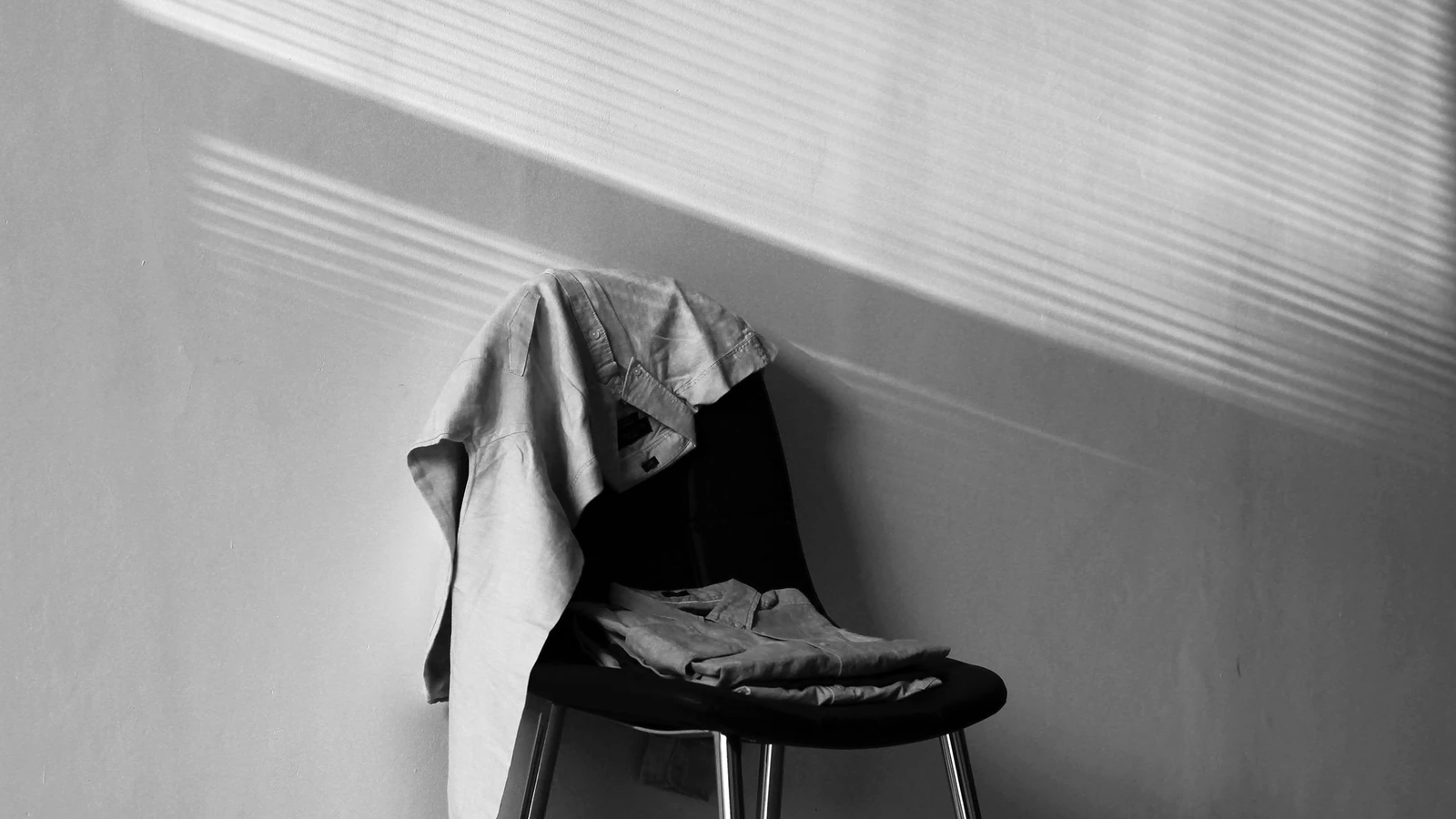 Un'immagine monocromatica che mostra una sedia coperta da vestiti sotto una luce e ombre drammatiche.