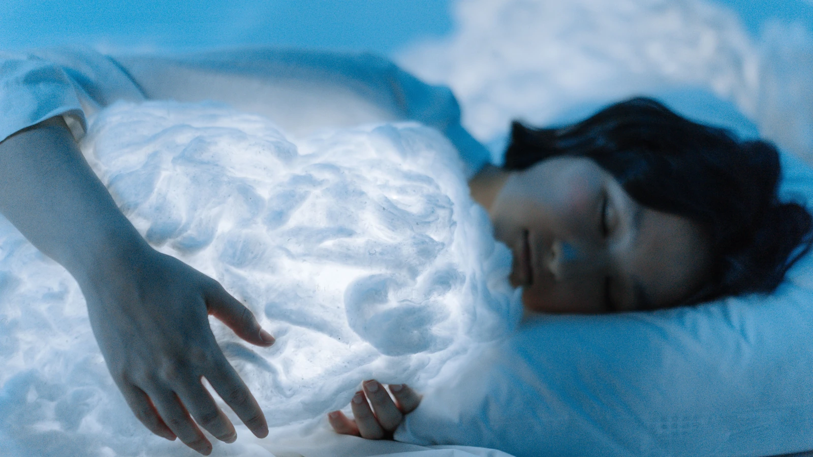 Una donna dorme serenamente abbracciando un cuscino a forma di nuvola luminosa, evocando tranquillità e stati da sogno.