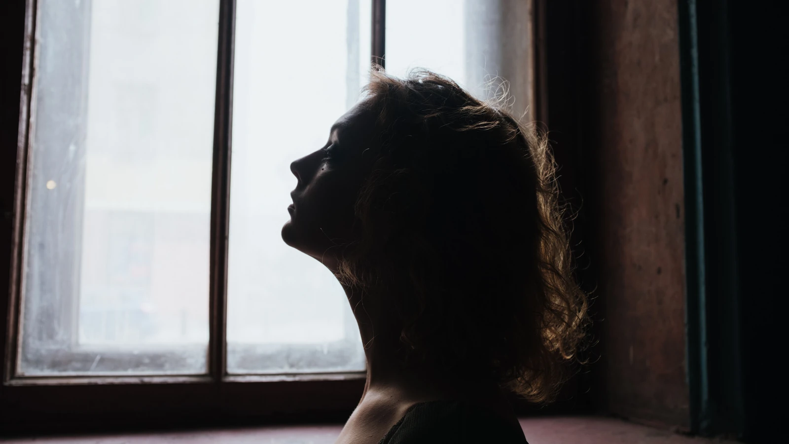 Silhouette di una donna con capelli ricci che guarda fuori da una finestra, creando un'atmosfera drammatica e contemplativa all'interno.