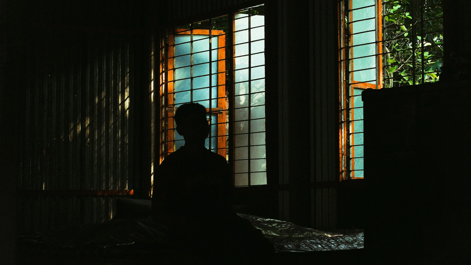 Silhouette di una persona seduta accanto a una finestra illuminata in una stanza buia, atmosfera suggestiva.