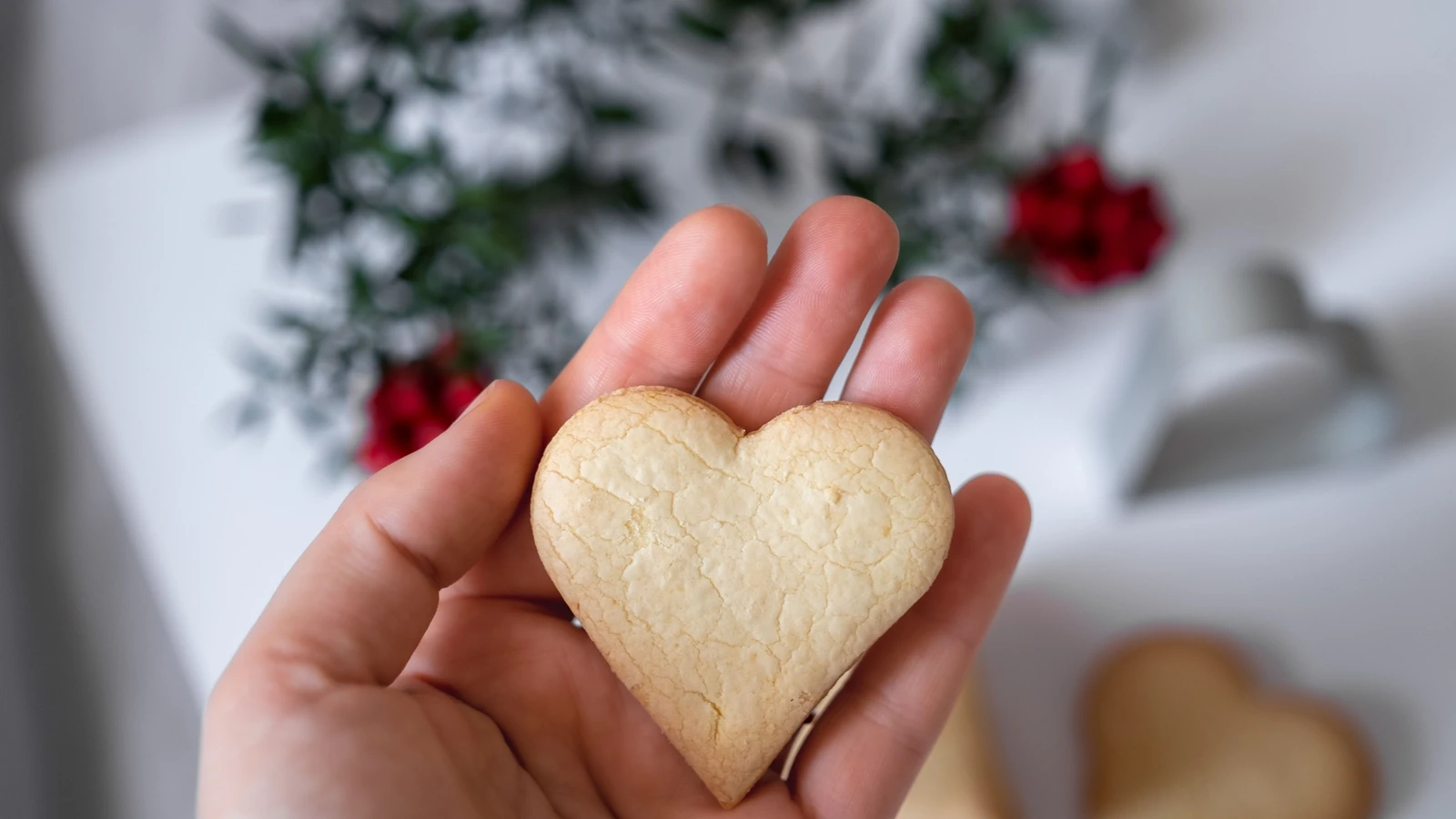 Una mano tiene un biscotto a forma di cuore con uno sfondo floreale sfocato, trasmettendo calore e affetto.