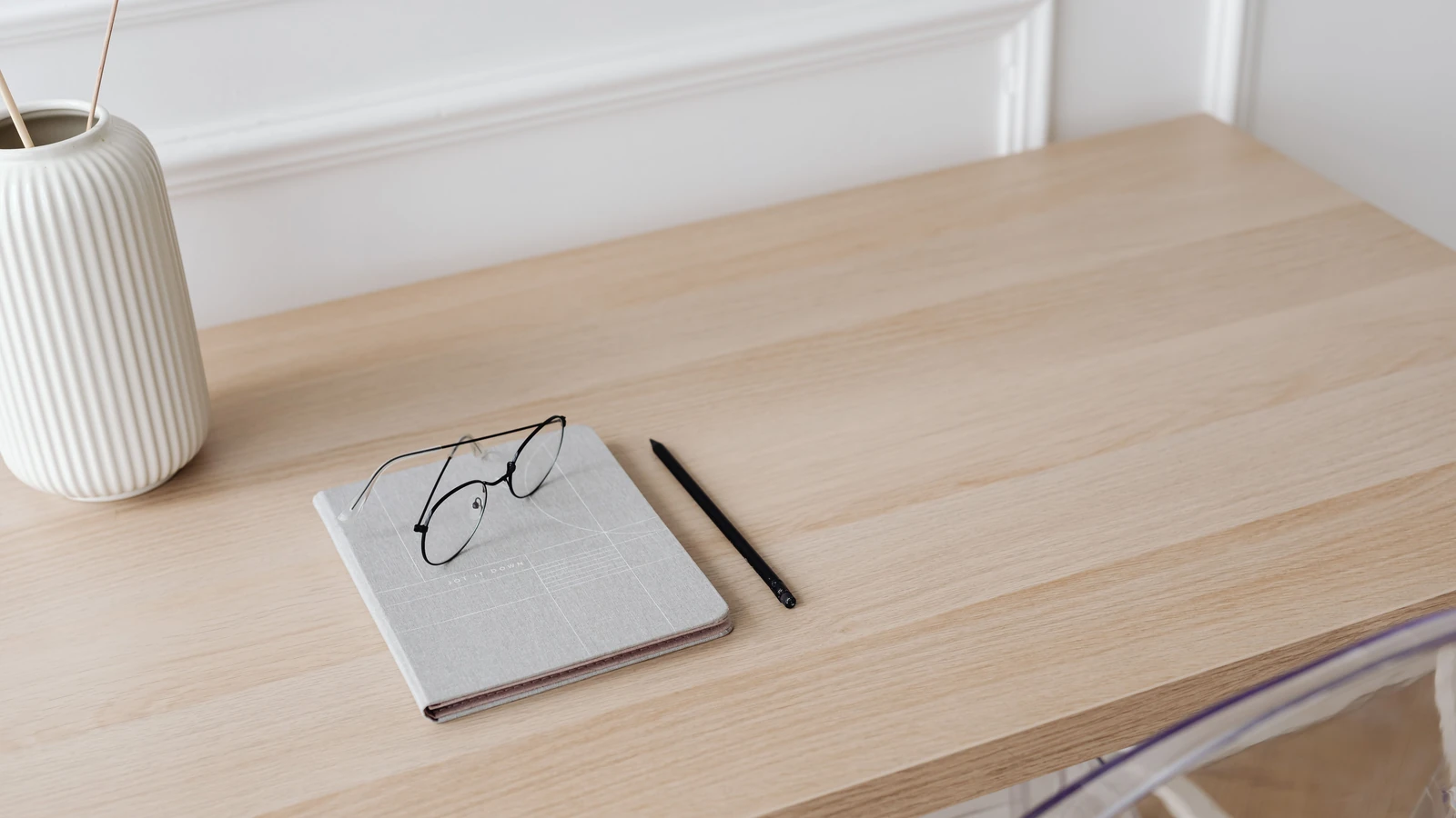 Uno spazio di lavoro minimalista con un quaderno, occhiali, penna e un vaso decorativo su una scrivania in legno.