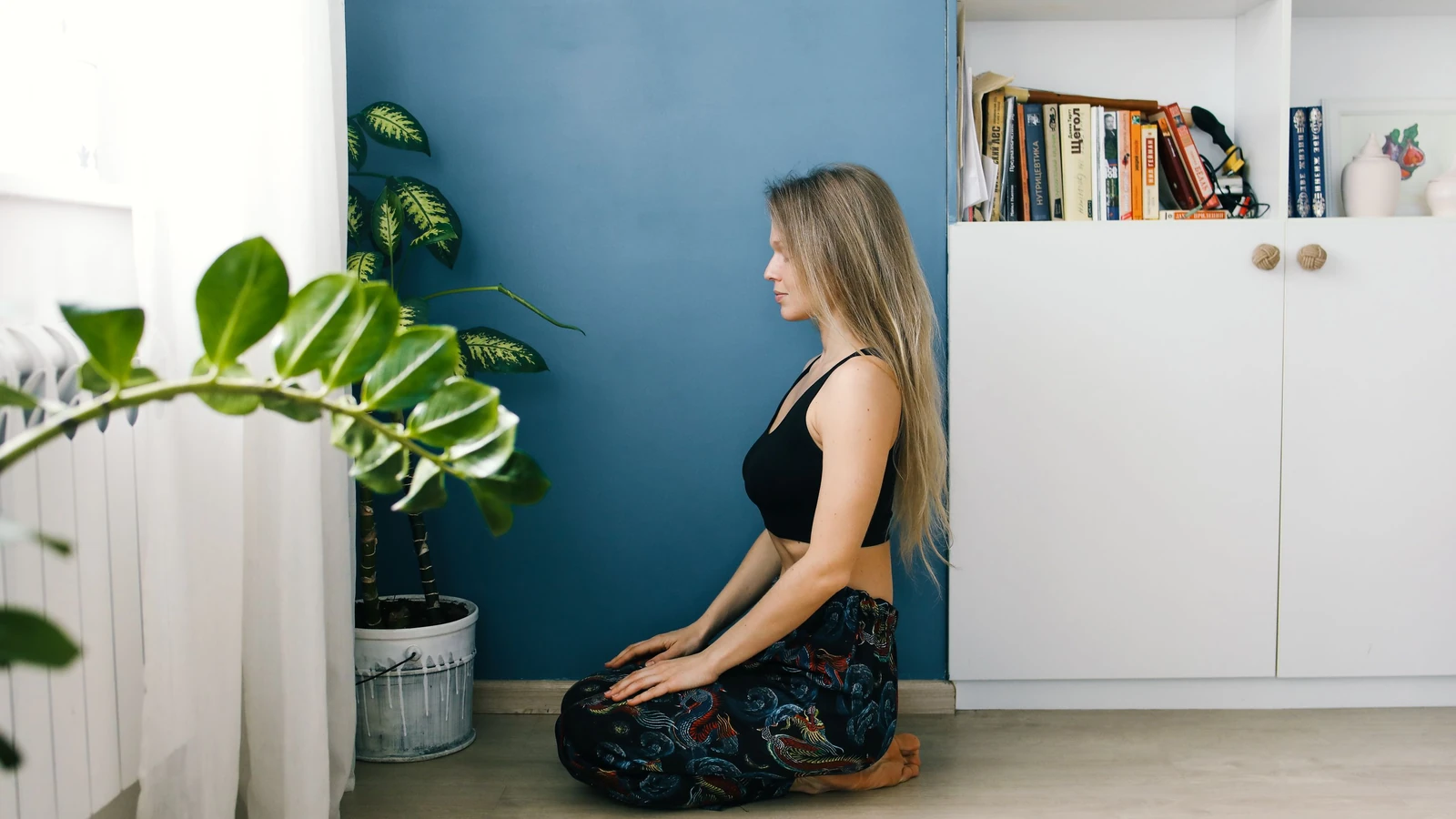 Donna serena che pratica meditazione in interni accanto a piante da appartamento e a una libreria.