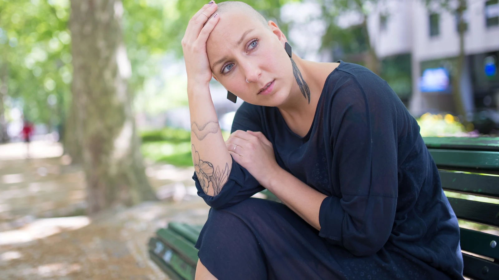 Donna calva con tattoo riflette su una panchina in un parco lussureggiante e soleggiato, simboleggiando la guarigione e l'introspezione.
