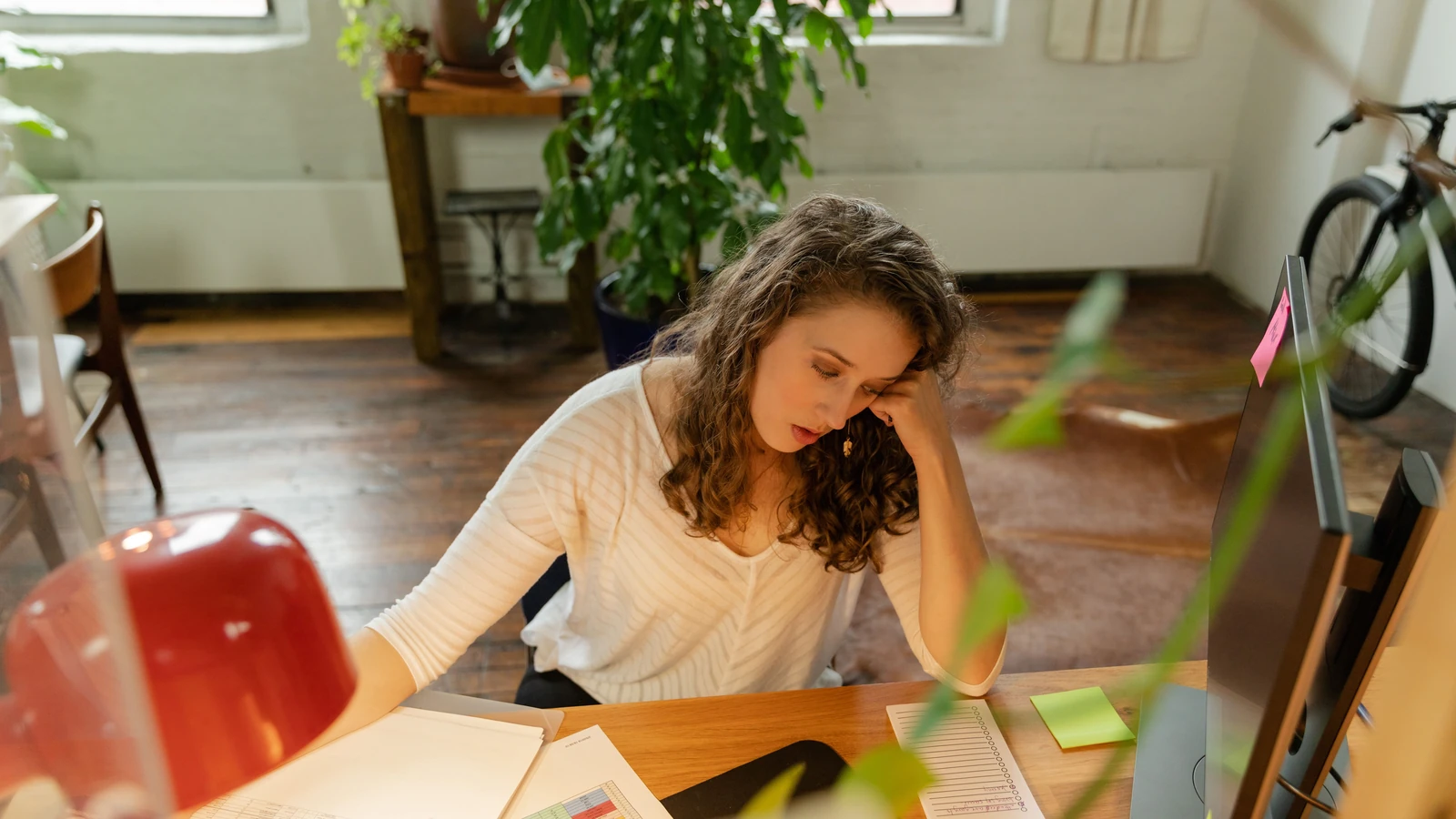 Una donna è seduta a una scrivania di legno, appare stanca mentre lavora in ufficio con post-it e un monitor.