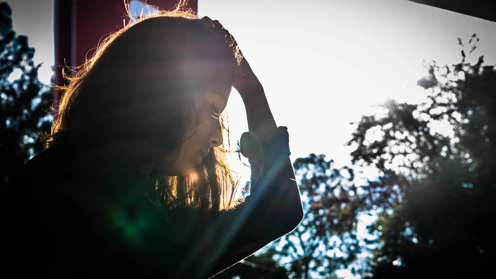 Silhouette di una donna che si tocca la testa, circondata dalla natura e dalla luce del sole. — Austin Guevara – Pexels