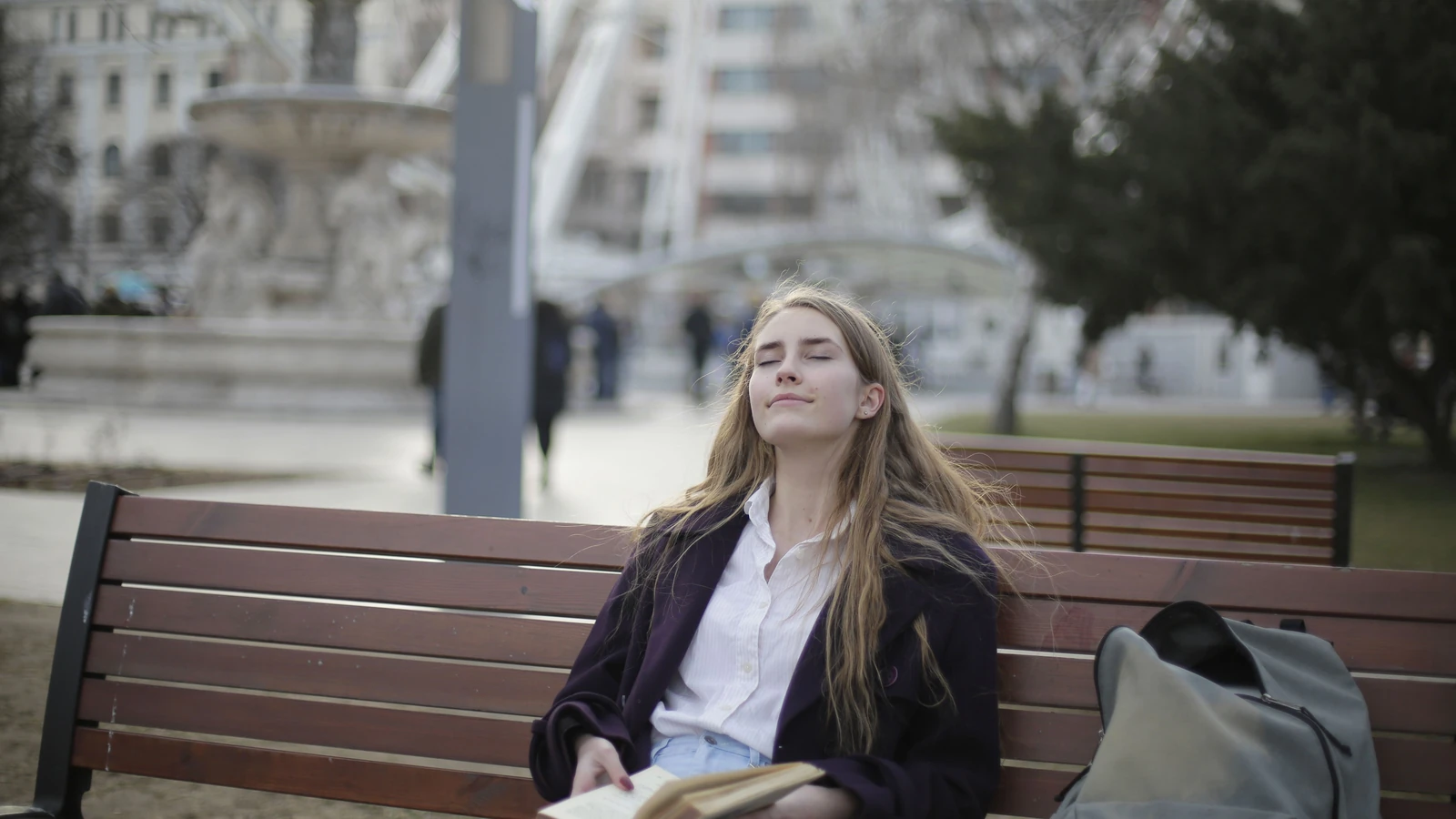 Donna che si gode un momento di relax su una panchina del parco con gli occhi chiusi e un libro in mano.