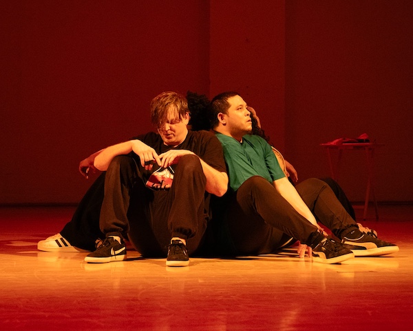 5 male dancers sit on the stage in red light, backs to each other, looking tired.