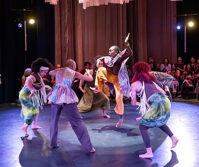 A photo of dancers in colorful Senegalese print costumes in a circle with Oluyinka mid jump in the air in the middle.