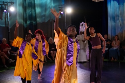 Ensemble of dancers on stage in shiny golden choir robes and purple sashes with their hands up grabbing the air with purpose.