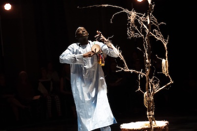 Massamba Diop playing Tama next to a sculpture of a tree in a black theater.