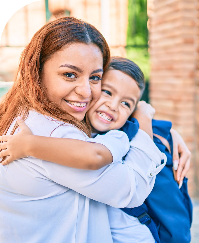 Smiling woman hugging a happy young boy wearing a blue backpack outdoors.