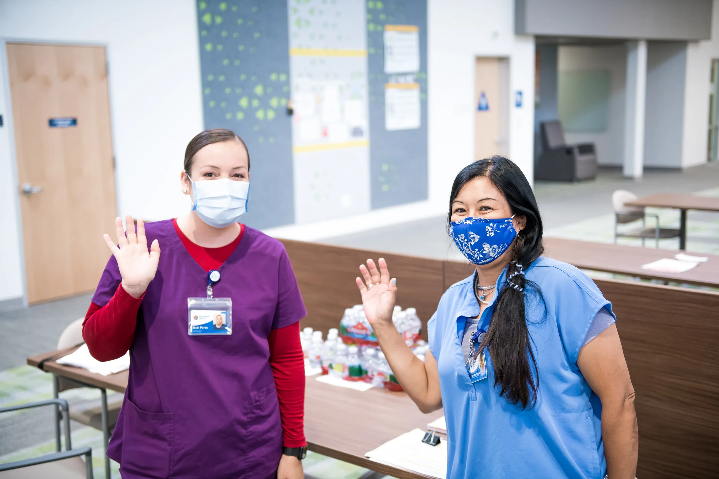 Two healthcare workers wearing face masks and scrubs waving inside a medical facility.