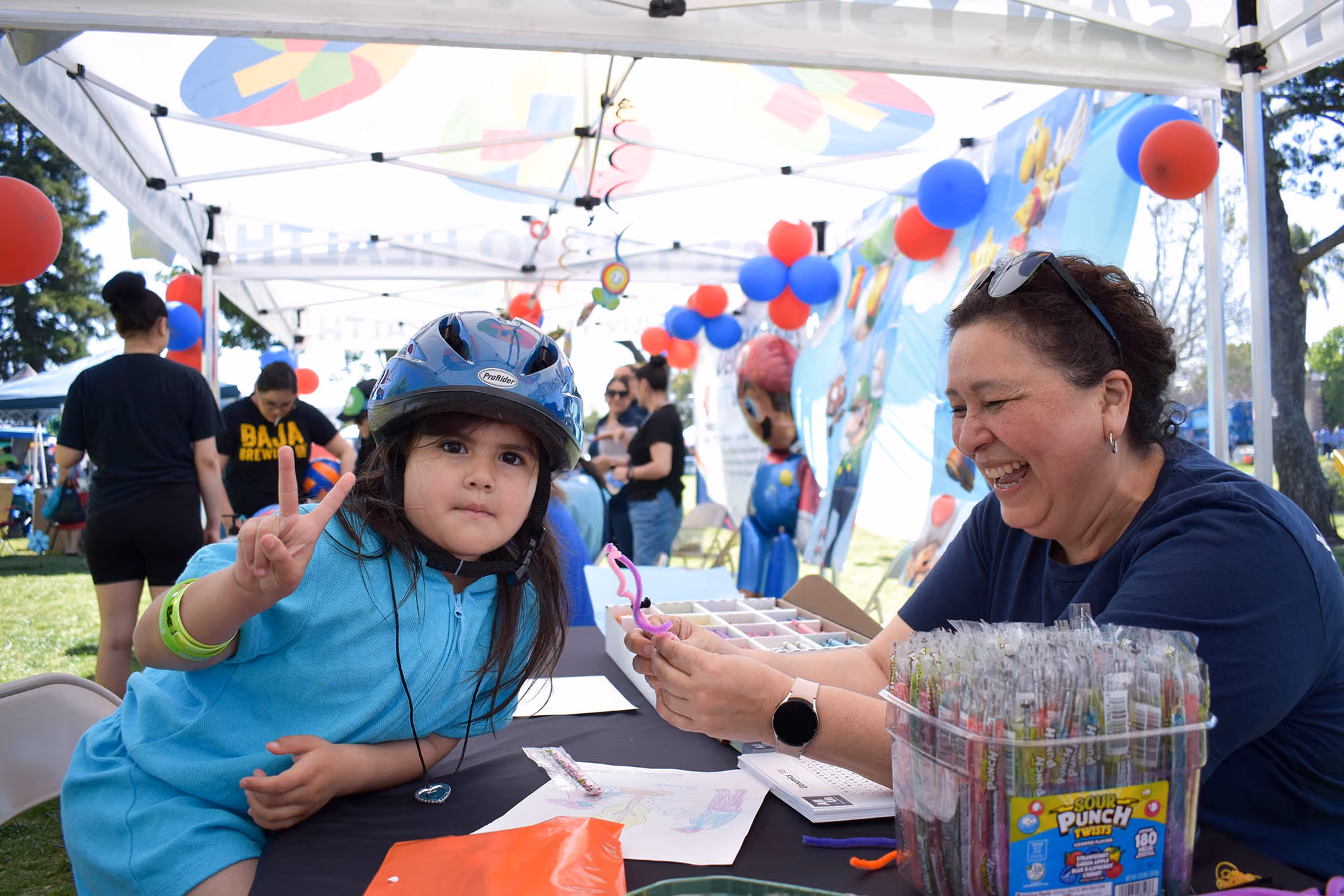 Una joven con casco azul y sudadera con capucha hace el signo de la paz mientras está sentada a una mesa con una mujer sonriente que sostiene materiales de manualidades bajo una carpa decorada.