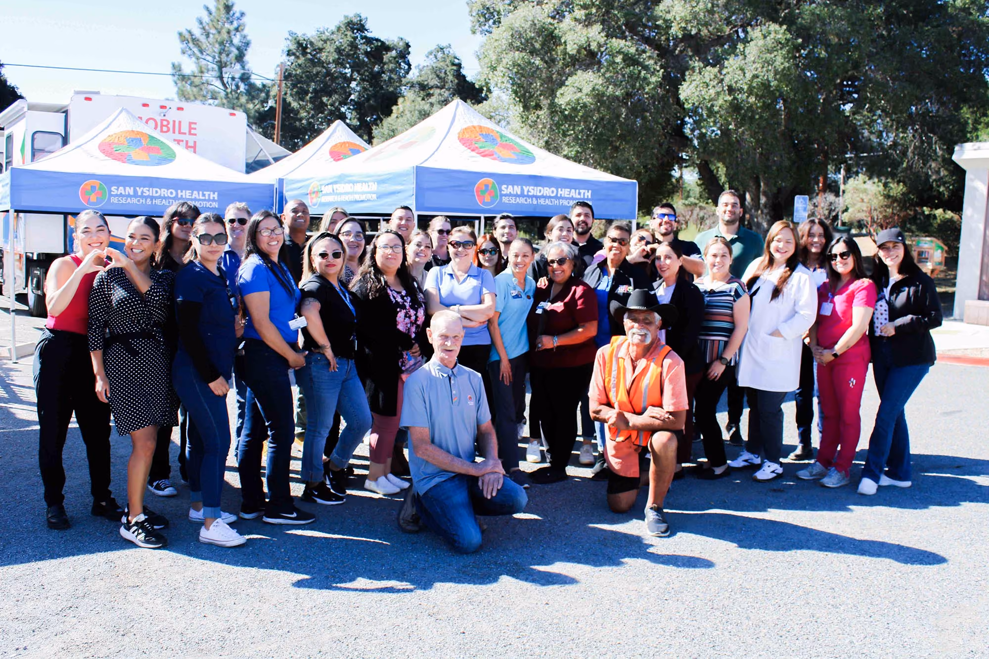 Grupo de trabajadores sanitarios y comunitarios de diversa procedencia posando al aire libre frente a las tiendas de campaña San Ysidro en un día soleado.