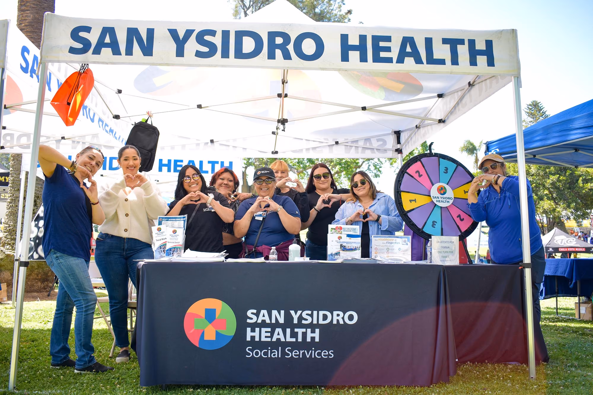 Grupo de nueve personas sonriendo y haciendo corazones con las manos bajo una carpa de Servicios Sociales San Ysidro en un evento al aire libre.