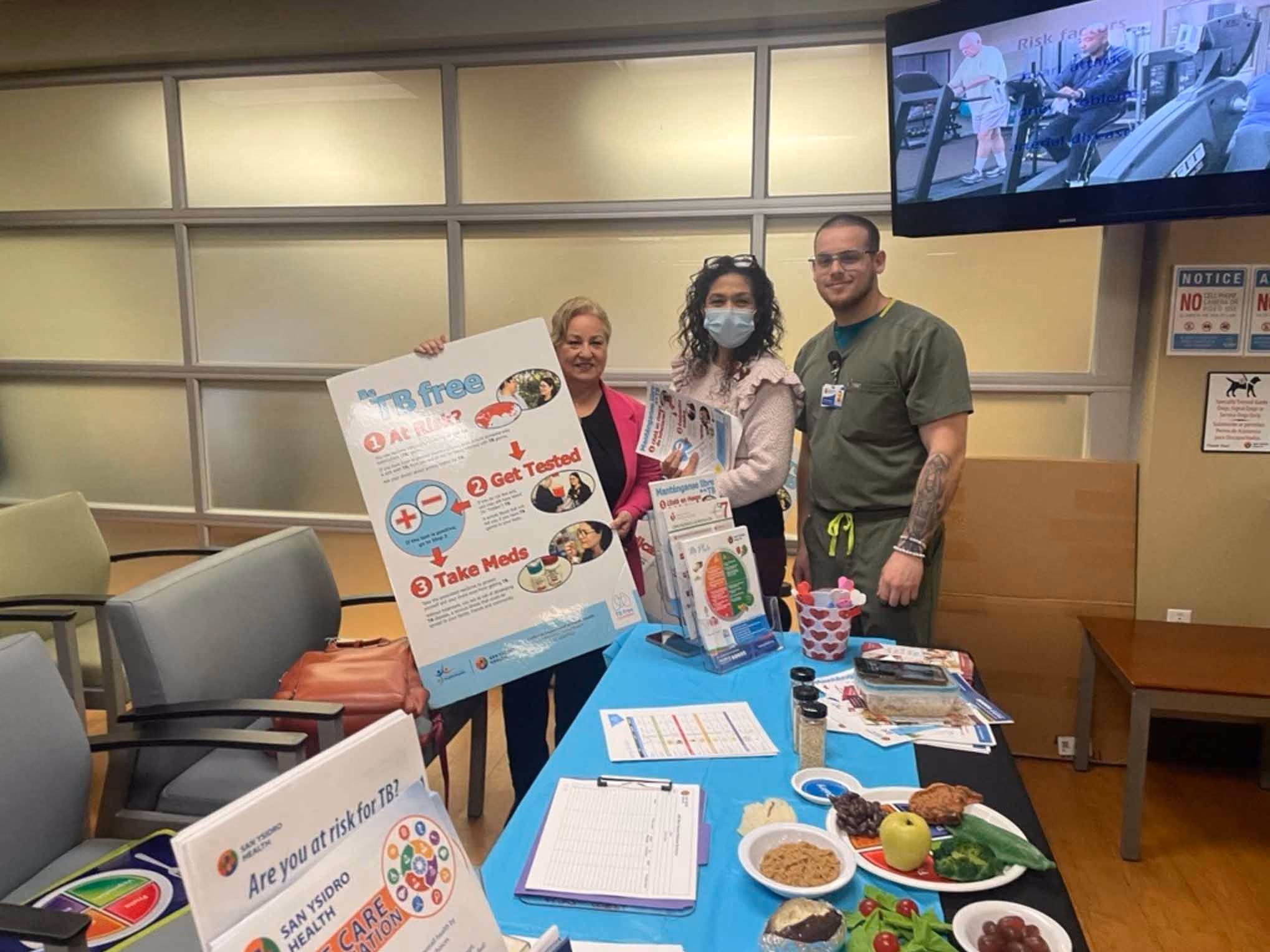 Three healthcare workers stand behind a table with TB awareness materials and healthy food samples in a clinic waiting area.