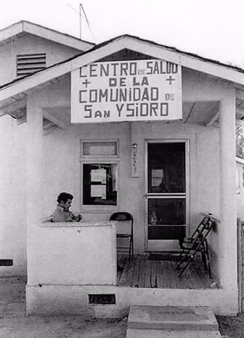 Fotografía en blanco y negro del porche de un pequeño centro de salud comunitario con un hombre sentado, un cartel que dice «Centro de Salud de la Comunidad de San Ysidro y dos sillas vacías.