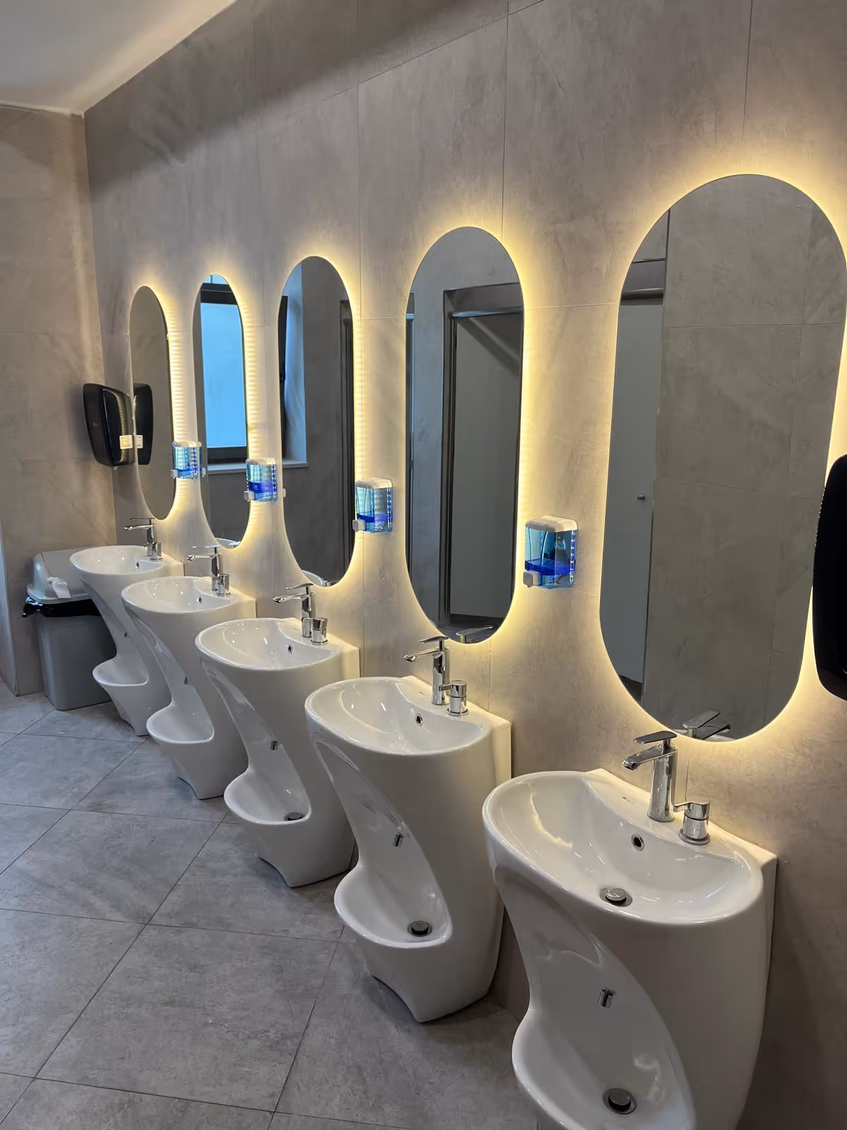 Row of five modern white sinks each with oval backlit mirrors and blue soap dispensers in a restroom.