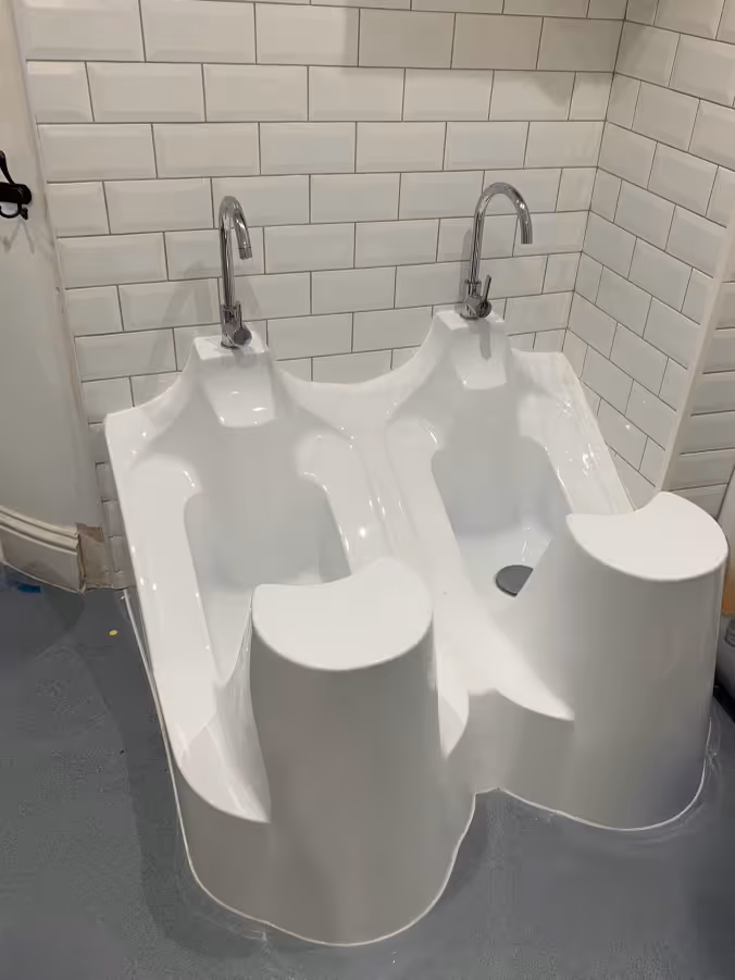 Dual white foot baths with chrome faucets set against white tiled wall on gray floor.