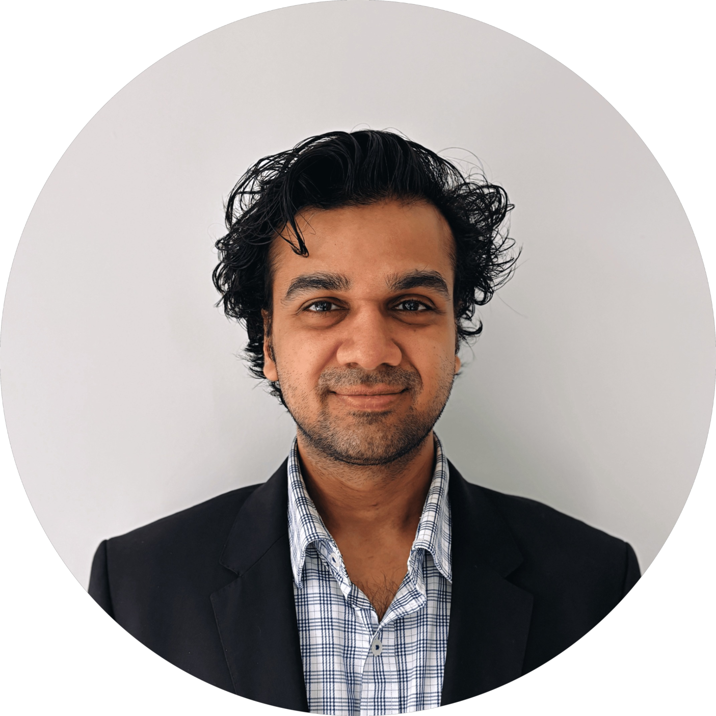 Man with curly dark hair wearing a black blazer and checkered shirt, smiling and facing the camera against a plain white background.
