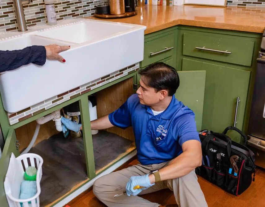 Stan's Heating, Plumbing, and Electrical technician working under a kitchen sink, with a customer pointing at the area of concern.