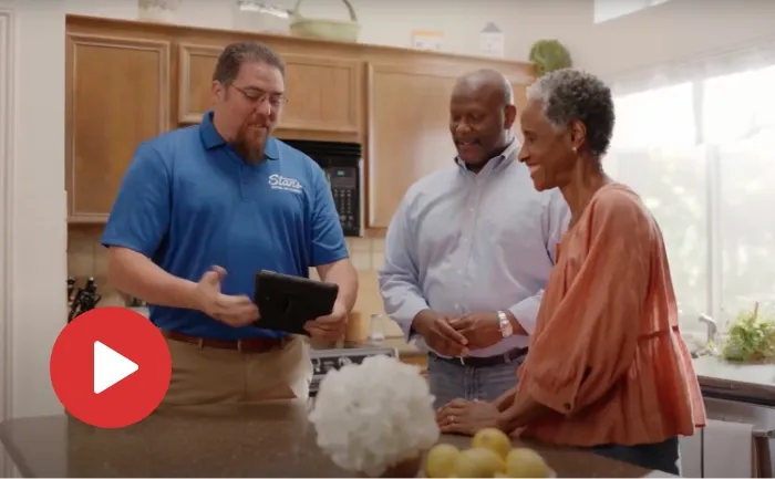Stan's Heating, Plumbing, and Electrical technician showing a couple information on a tablet in a kitchen, with a play button icon indicating a video.