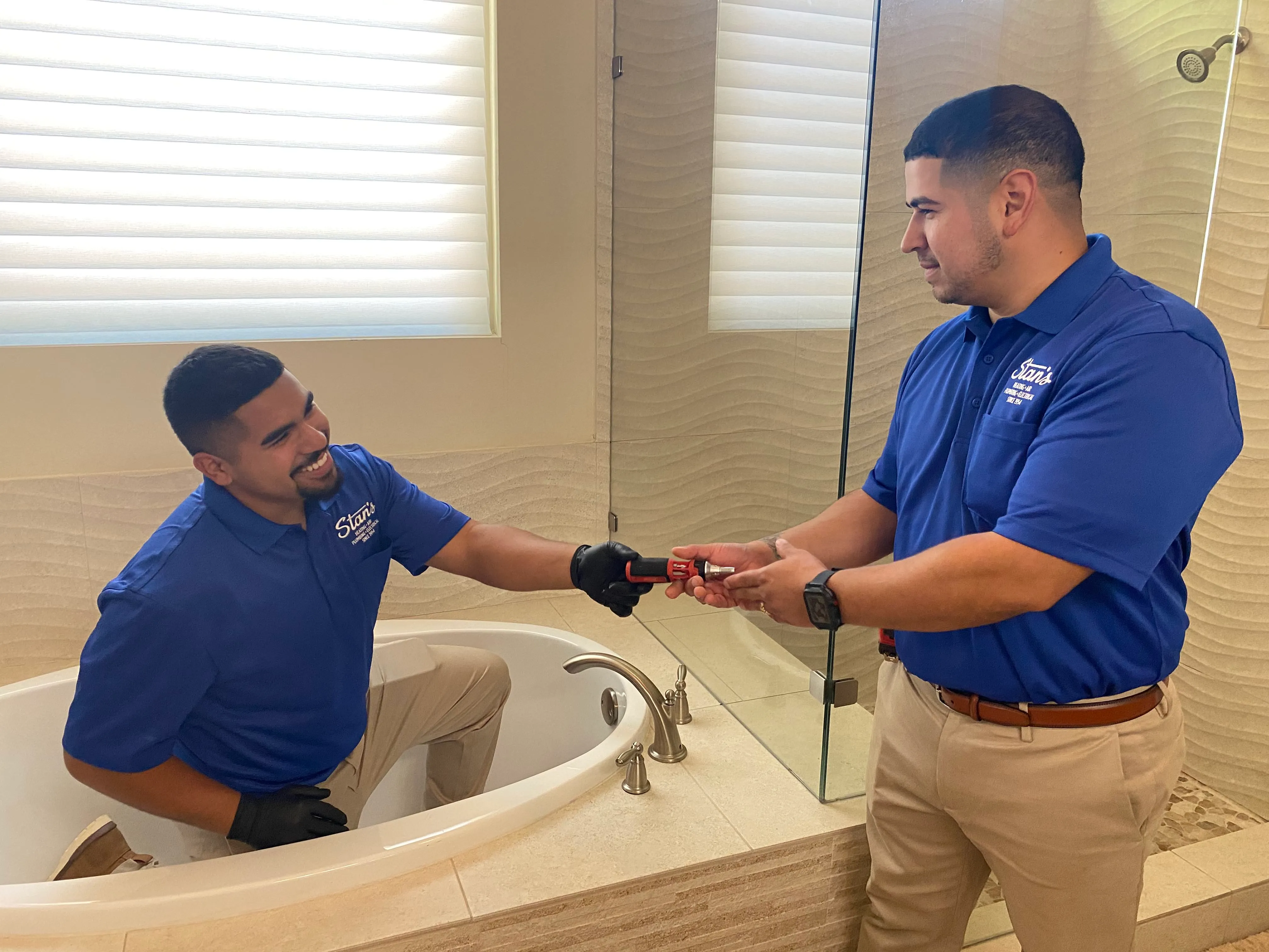 Two technicians in blue Stan's Heating, Air, and Plumbing shirts working together in a bathroom, one handing a tool to the other who is kneeling in a bathtub.