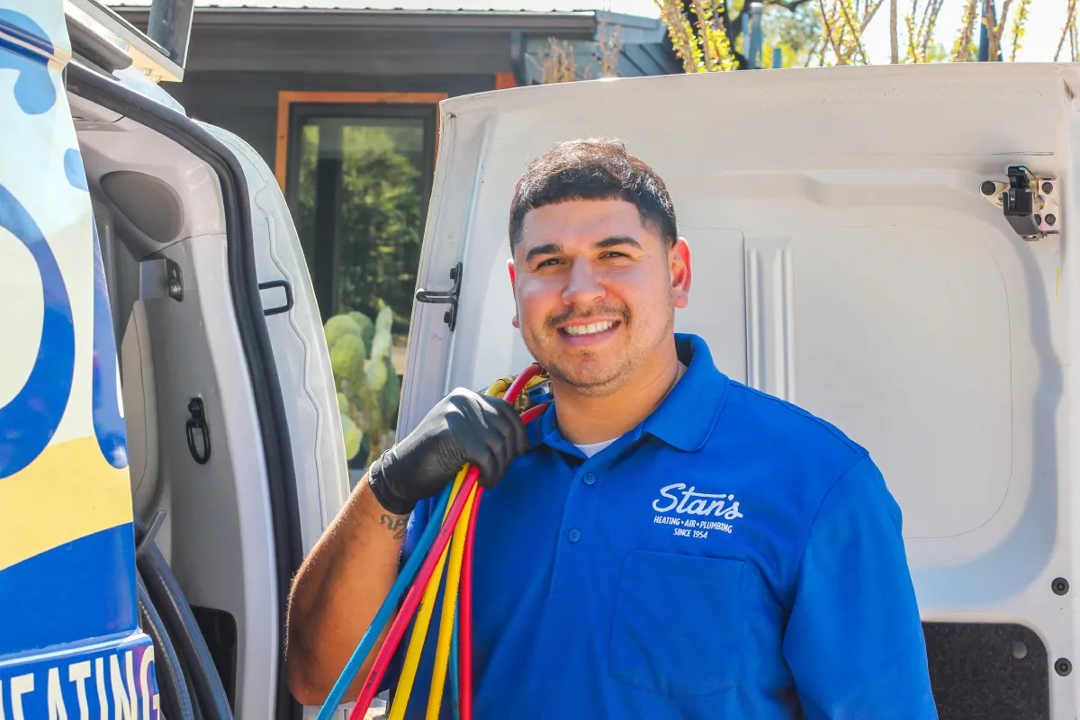 A smiling technician wearing a blue Stan's Heating, Air, and Plumbing shirt, holding colorful tubing and standing by the open door of a service van.