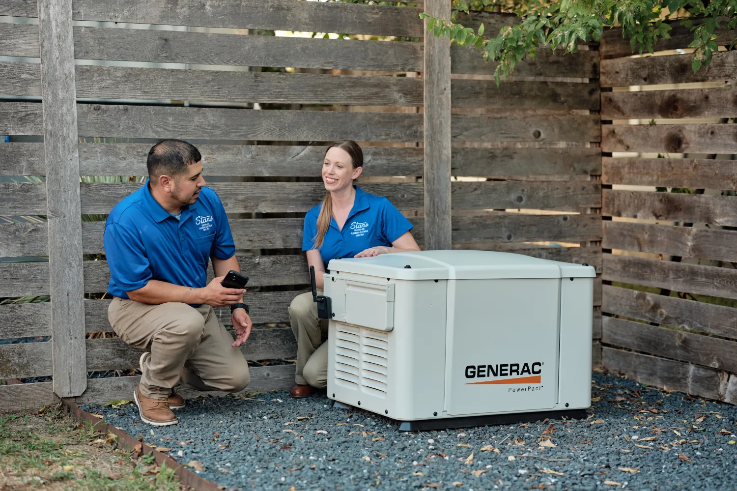 Two technicians from Stan's Heating, Air, Plumbing, and Electrical are discussing a Generac generator installed beside a wooden fence. One technician is holding a smartphone while the other is explaining something about the generator.