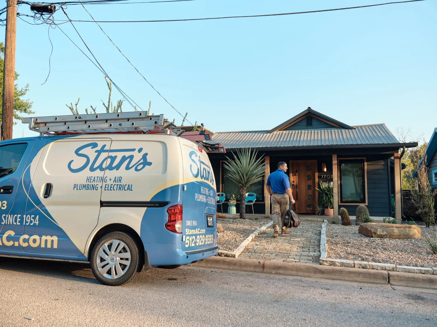 A technician from Stan's Heating, Air, Plumbing, and Electrical is walking towards a modern house with tools in hand. The company van is parked nearby.